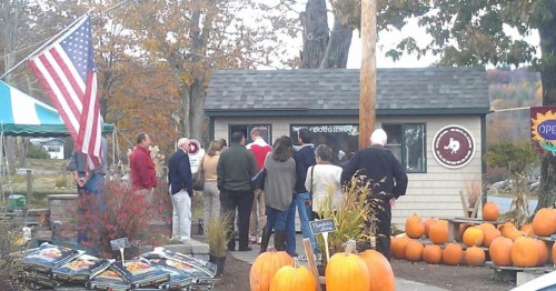 A group of people gathers outside a small building decorated with pumpkins and an American flag.