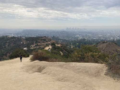 A person walks along a dirt path overlooking a hazy cityscape with hills in the foreground.