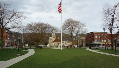 A park with a flagpole, American flag, trees, and buildings in the background under a cloudy sky.