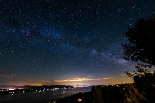 A starry night sky over a calm lake, with the Milky Way visible and distant lights along the shoreline.