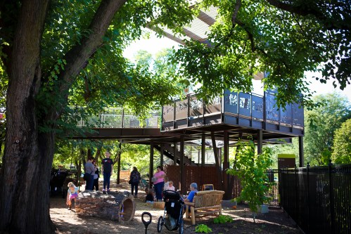 A playground with a treehouse, surrounded by trees, where children and adults enjoy outdoor activities.