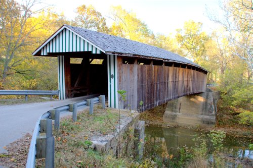 A wooden covered bridge with a sloped roof spans a calm stream, surrounded by autumn foliage and a gravel path.