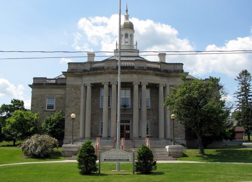 Historic building with columns, a clock tower, and a sign in front, surrounded by trees and a blue sky.