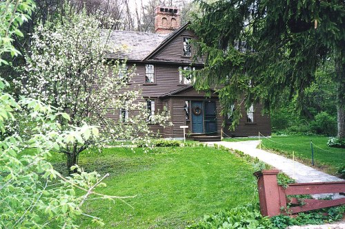 A brown wooden house surrounded by greenery and blooming trees, with a path leading to the front door.