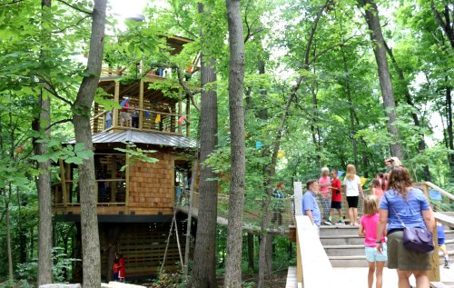 A treehouse surrounded by trees, with children playing and adults nearby on a wooden path.