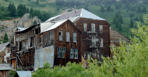 Abandoned wooden building with multiple stories, surrounded by greenery and mountains in the background.