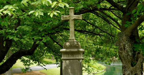 A weathered stone cross stands among lush green trees by a serene pond.