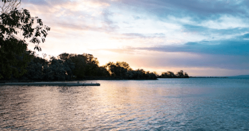 A serene lake at sunset, with trees lining the shore and colorful clouds reflecting on the water's surface.