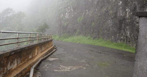 A misty road winding alongside a rocky cliff, with lush greenery and a metal railing on one side.