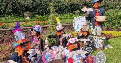 A whimsical tea party scene with people in colorful hats and orange wigs, enjoying tea in a vibrant garden setting.