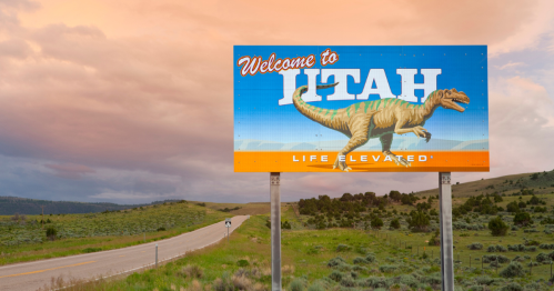 A colorful "Welcome to Utah" sign featuring a dinosaur, set against a scenic landscape and cloudy sky.