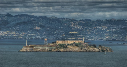 Alcatraz Island surrounded by water, with a cloudy sky and distant hills in the background.