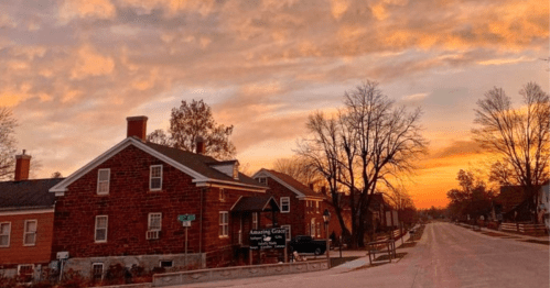A picturesque sunset over a quiet street lined with charming houses and trees, casting warm colors in the sky.