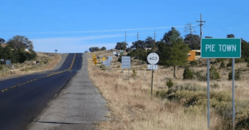 A road sign for Pie Town alongside a highway with various directional signs and a clear blue sky.