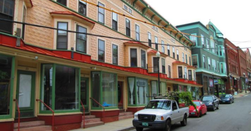 A street view of colorful, historic buildings with shops on the ground floor and parked cars along the curb.