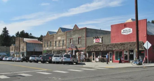 A quaint small-town street with historic buildings and a grocery store, lined with parked cars under a clear blue sky.