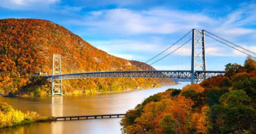 A scenic view of a bridge over a river, surrounded by vibrant autumn foliage and rolling hills under a blue sky.