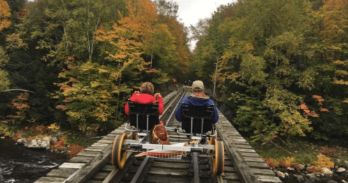 Two people ride a rail bike on a wooden track surrounded by colorful autumn trees.