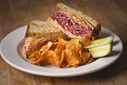 A plate with a stacked sandwich filled with meat and cheese, served with crispy chips and pickle slices on the side.