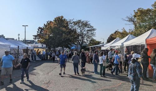 A bustling outdoor market with people browsing vendor tents and enjoying the sunny day. Dogs are present among the crowd.