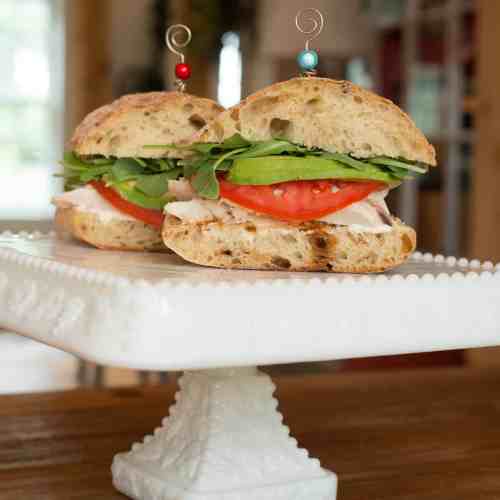 Two sandwich halves on a white cake stand, featuring turkey, tomato, and greens, topped with decorative skewers.