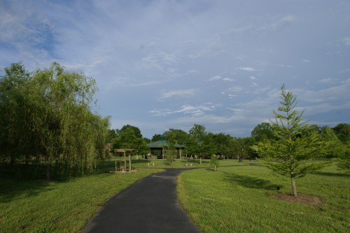 A winding path through a green park with trees and a gazebo under a partly cloudy sky.
