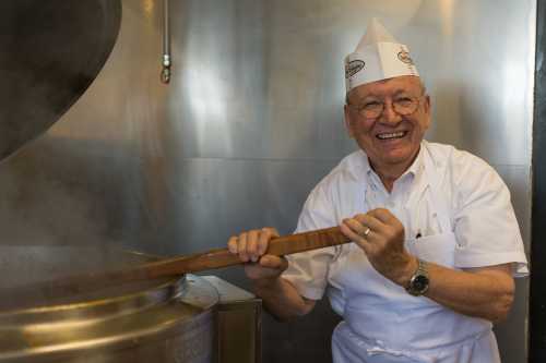 A smiling chef in a white uniform and hat stirs a large pot in a kitchen filled with steam.