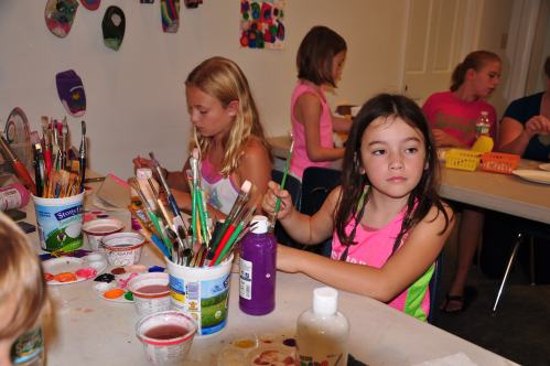 Children engaged in an art activity, painting with brushes and colorful paints at a table in a creative space.