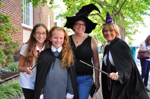 Four women and girls dressed in witch costumes, holding wands, smiling outdoors with trees in the background.