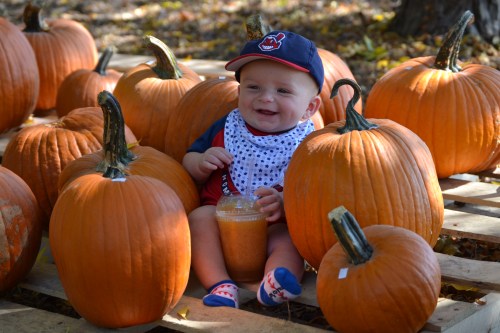 A smiling baby sits among large pumpkins, wearing a cap and a bib, holding a drink in a fall setting.