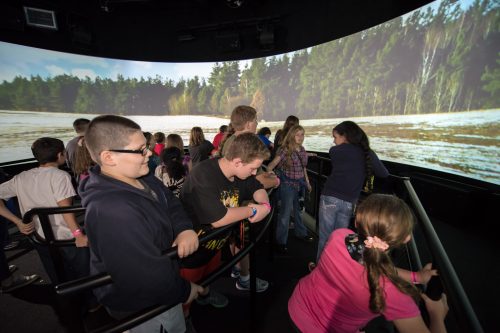 A group of children stands inside a dark room, watching a large immersive nature video on a curved screen.