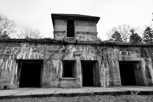 A weathered, abandoned building with a tower and three dark doorways, surrounded by bare trees in black and white.
