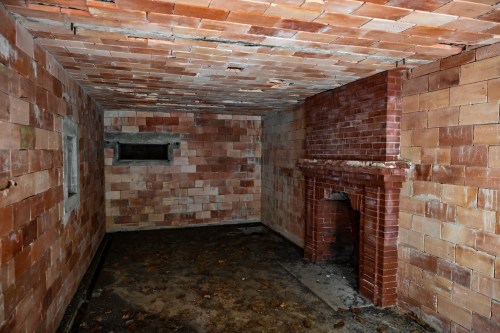 An empty, dimly lit room with brick walls and a brick fireplace, featuring a dirty floor and a small window.
