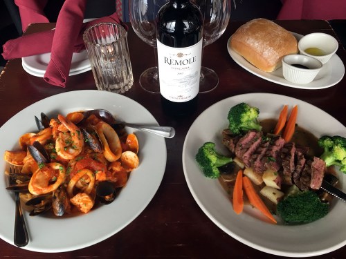Two plates of food: one with seafood pasta and the other with steak, broccoli, and carrots, accompanied by wine and bread.