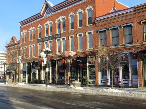 Historic brick buildings line a snowy street, featuring shops and cafes with colorful signage.