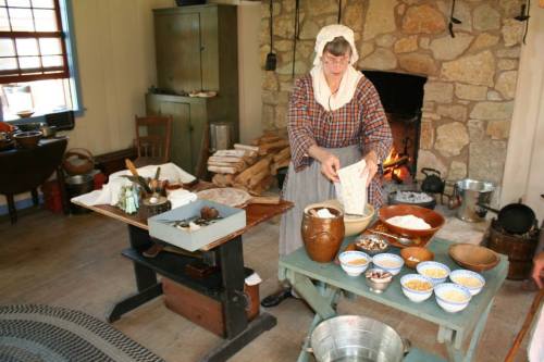 A woman in historical attire prepares food in a rustic kitchen with a stone fireplace and various bowls of ingredients.