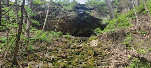 A rocky landscape with a large overhang, surrounded by trees and greenery, leading to a forested area.