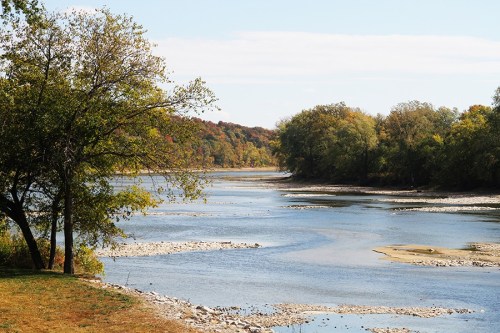 A serene river scene with trees lining the banks and colorful autumn foliage in the background under a clear blue sky.