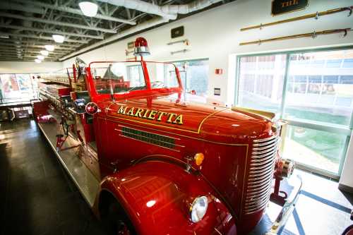 A vintage red fire truck with "MARIETTA" written on the side, displayed indoors with large windows in the background.