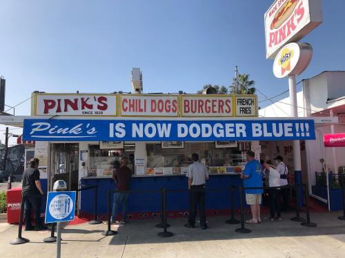 A busy hot dog stand with a sign reading "Pink's is now Dodger Blue!" and people ordering food.
