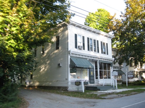 A white two-story building with green awnings, surrounded by trees and a sidewalk, in a quiet neighborhood.