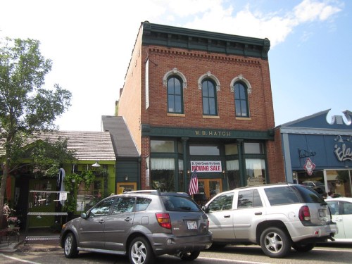 Historic brick building with "W. B. Hatch" sign, featuring a large window and a "Going Sale" banner, surrounded by cars.