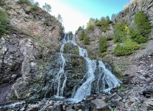 A cascading waterfall flows down rocky cliffs, surrounded by lush greenery and a clear blue sky.