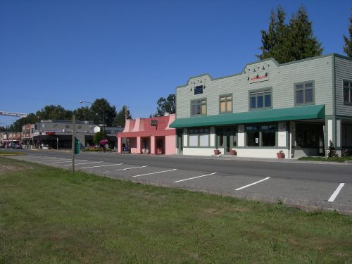 A row of small businesses with colorful storefronts along a quiet street under a clear blue sky.