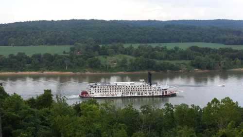 A large steamboat navigates a river surrounded by lush green hills and trees under a cloudy sky.