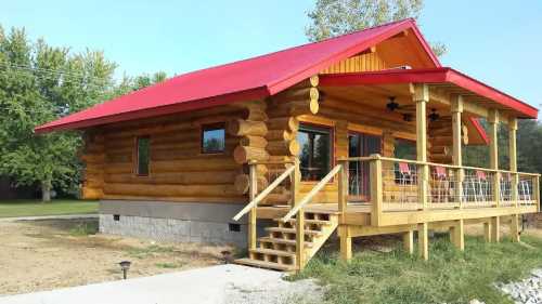 A cozy log cabin with a red roof, wooden deck, and large windows, surrounded by greenery and a clear blue sky.