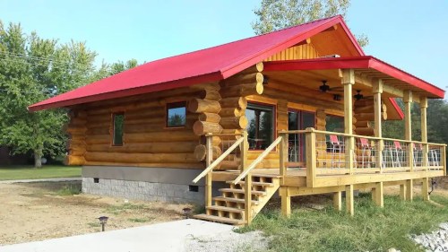 A cozy log cabin with a red roof, wooden deck, and large windows, surrounded by greenery and a clear blue sky.