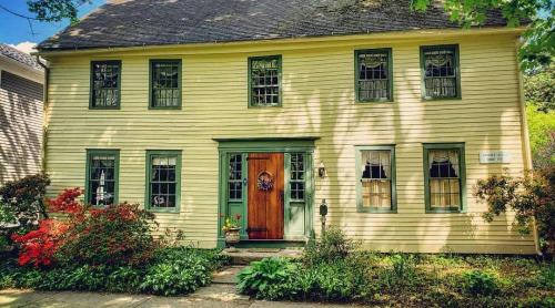 A charming yellow house with green trim, featuring a wooden front door and surrounded by lush greenery and flowers.