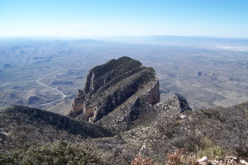 A panoramic view of a rugged mountain peak surrounded by vast, arid landscapes under a clear blue sky.