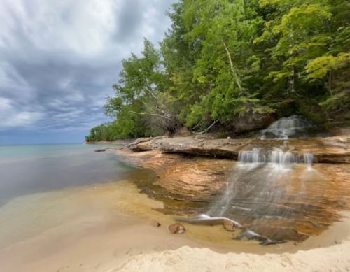 A serene beach scene with a gentle waterfall cascading over rocks, surrounded by lush green trees and a cloudy sky.